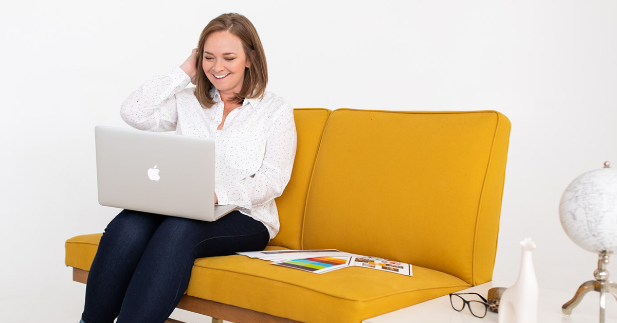 Photo of HeartSpark's founder, Lauren Atherton sitting on a vintage yellow couch. She is smiling while looking at her laptop and tucking her hair behind her ear.