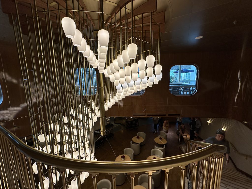 The lobby of "The Wake" restaurant aboard Virgin Voyages Valiant Lady ship, featuring a spiral art-deco inspired chandelier with encircled by a spiral staircase.