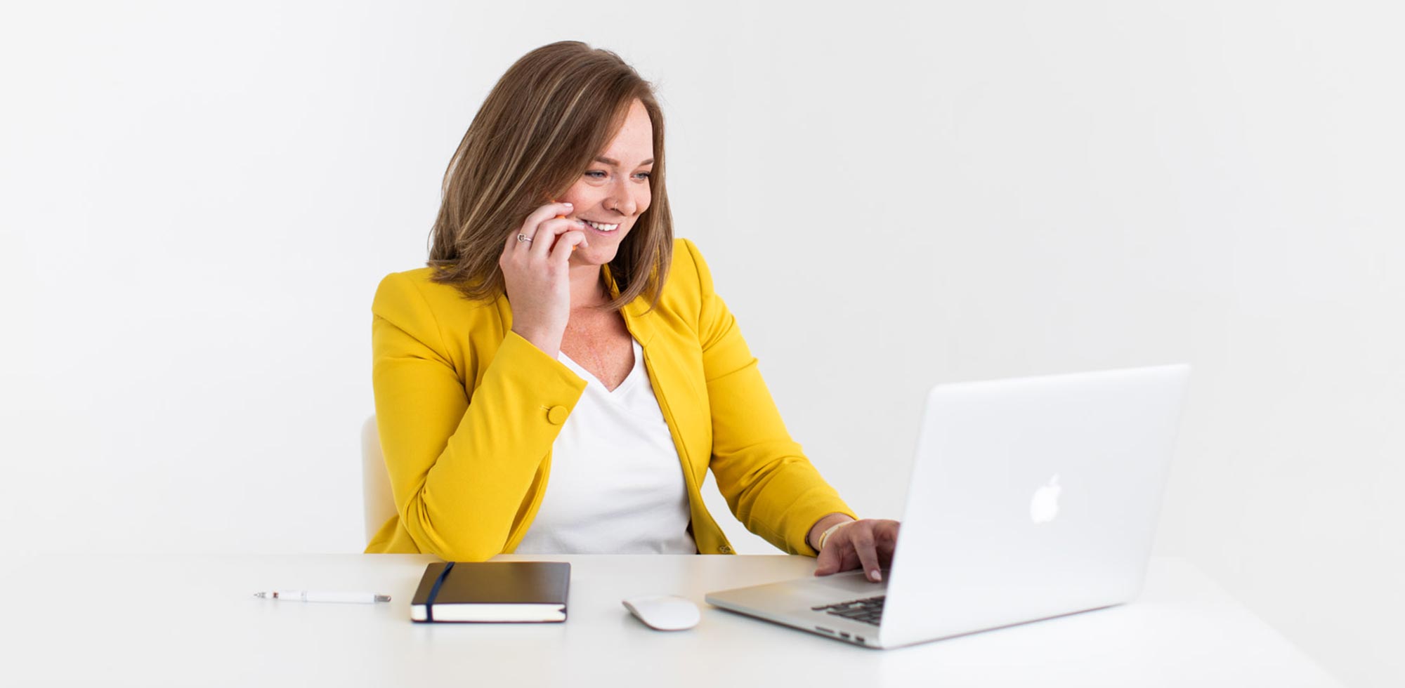 Woman in a yellow blazer smiling while sitting at a white desk and talking on the phone. She's typing notes on her laptop.